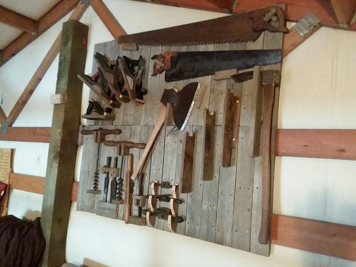 planes, spokeshaves, augers, crosscut saws, broadaxe, foot adze and other tools hanging on a rack