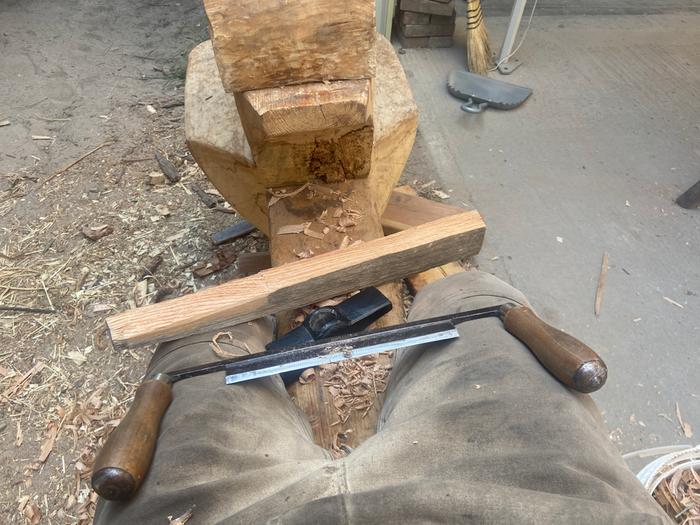 a drawknife sitting on a woman's legs at a shaving horse with a piece of wood