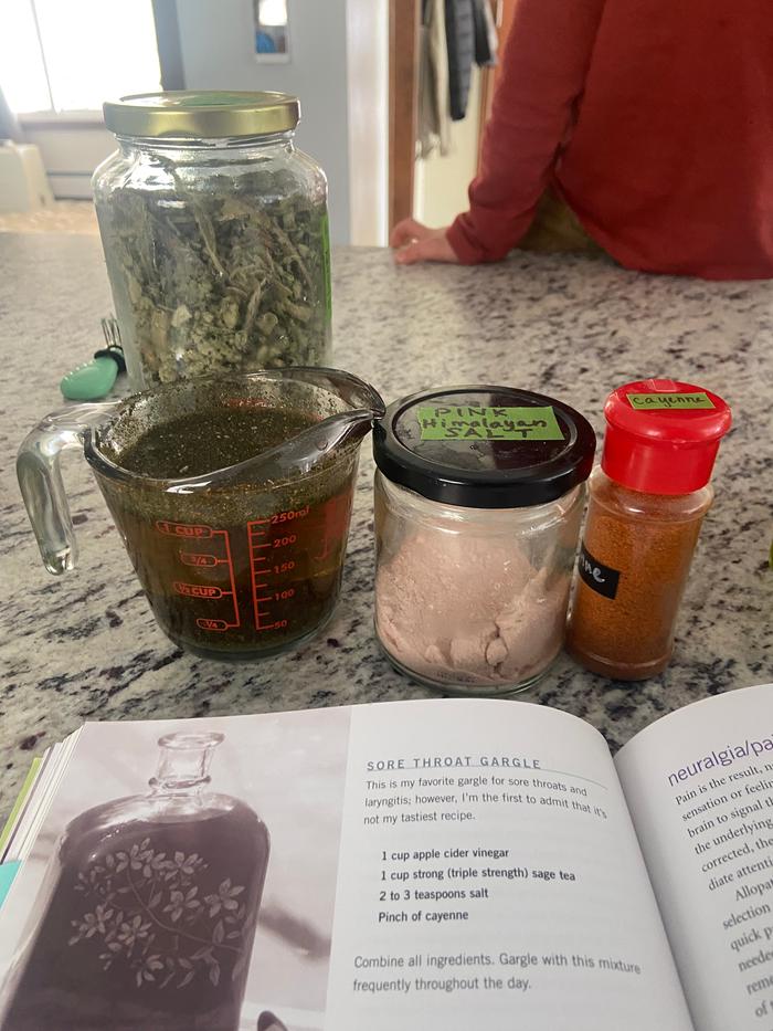 a sore throat gargle made from sage, vinegar, salt, and cayenne in a measuring cup with a glass jar of pink himalayan salt and a plastic jar of cayenne pepper on a stone counter in front of a book with the recipe