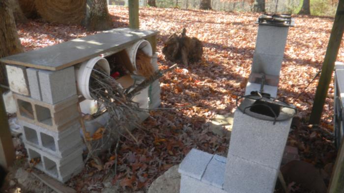 a workbench made from stacked cinder block with buckets of rocket fuel across from some rocket cookstoves in a pergola with a leaf covered yard behind it