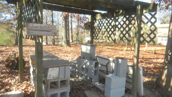 A pergola that has been converted to an outdoor kitchen by adding three rocket stoves and a workbench. The woodbunred sign says it is the 