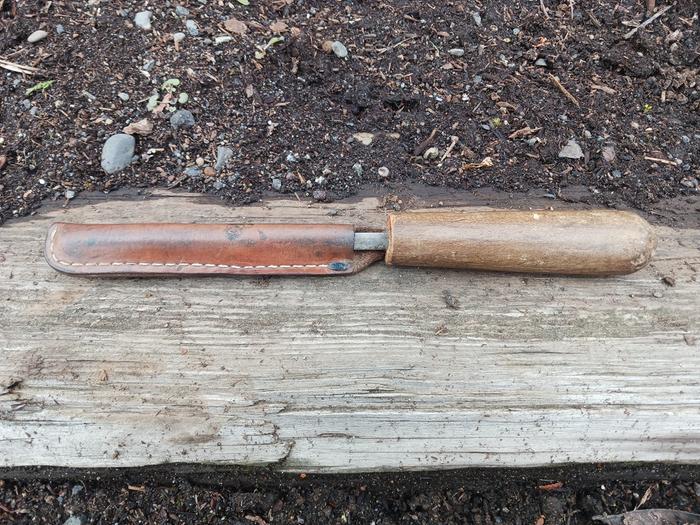 A knife steel in a leather sheath sitting on a board on dirt