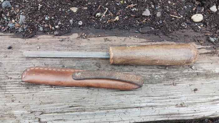 A knife steel, out of it's sheath, with a reclaimed wood handle, sitting on a board on the dirt