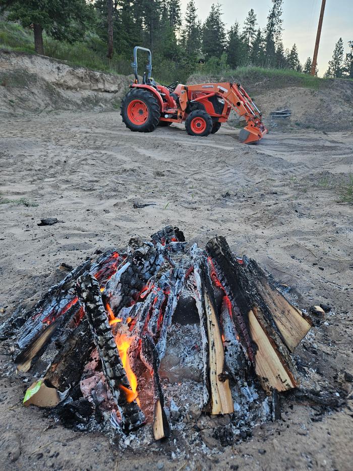 A bonfire in a sandlot to make bone sauce. A tractor sits at the far end of the sandlot