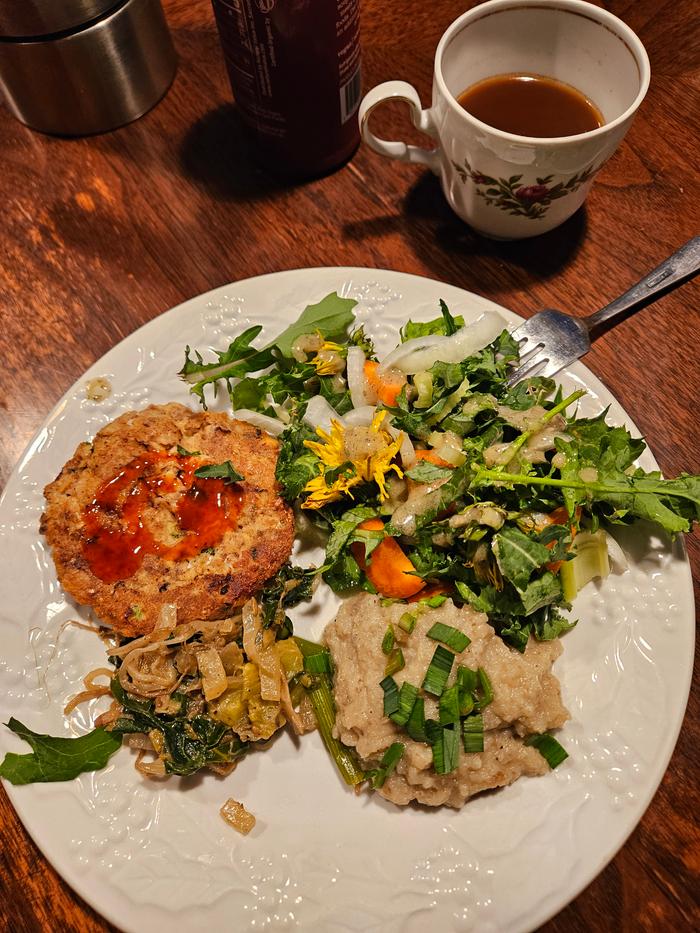 A delicious lunch of salmon patties, garden grown salad, mashed sunchokes and garlic on a white plat on a brown table with a cup of tea