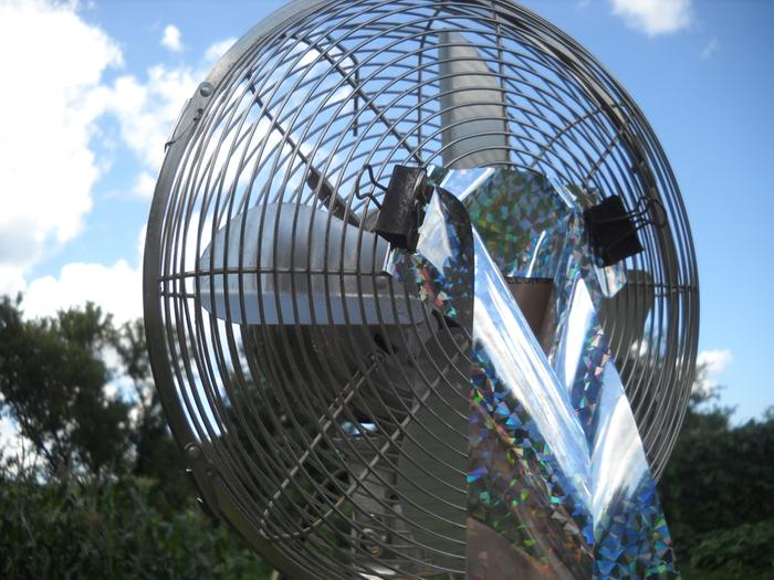 A metal fan with bird scaring streamers tied to it's cage outside