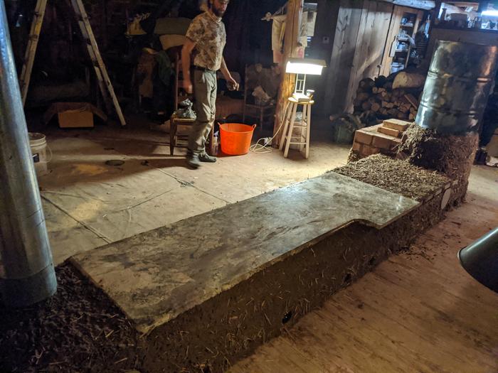 a slab from a granite countertop sits on top of the bench of a rocket masonry heater. A man stands in the background