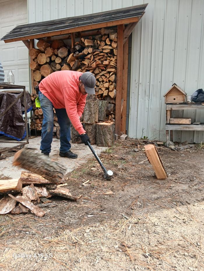 Esteban splits some firewood with a splitting maul in front of a full firewood rack