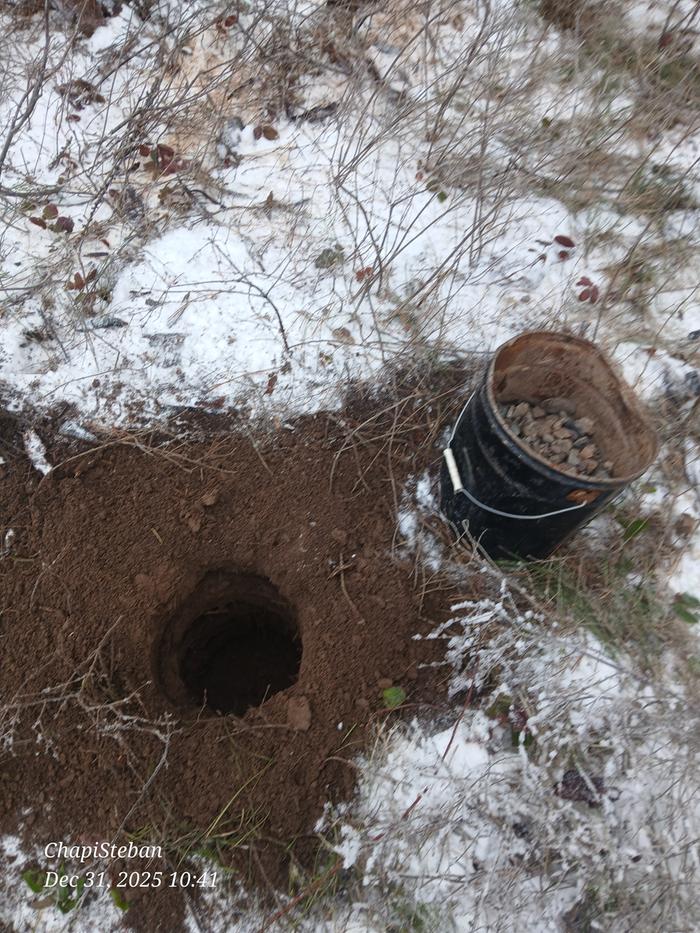 A hole for the upright support of a junkpole fence with a bucket of gravel next to it