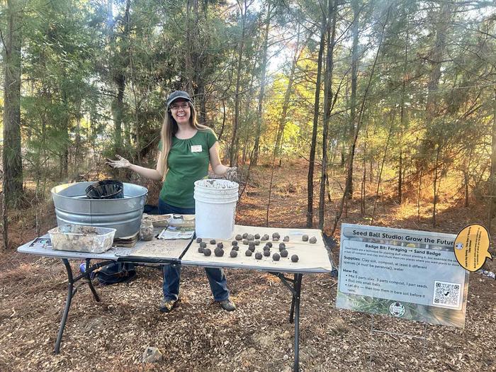 a woman in a green shirt with a hat shows people how to make seed balls at the Texas Permaculture Expo with evergreen trees in the background