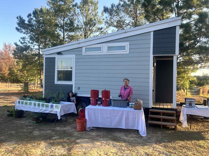Two women in front of a tiny house trailer show people how to recognize herbs