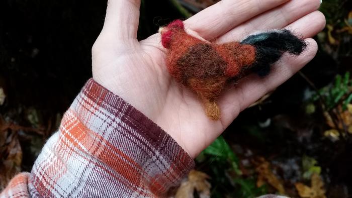 Needle-felted rooster held in a hand in the woods with a cat in the background